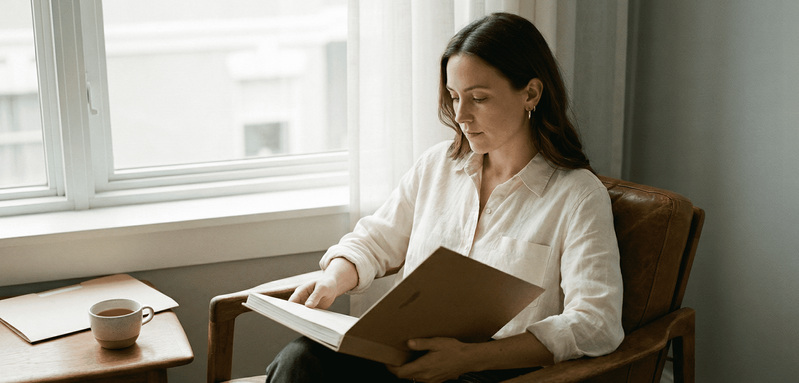 Woman in her late 30s sitting thoughtfully in a mid-century leather chair by a window, holding a closed portfolio, with soft diffused daylight illuminating her profile